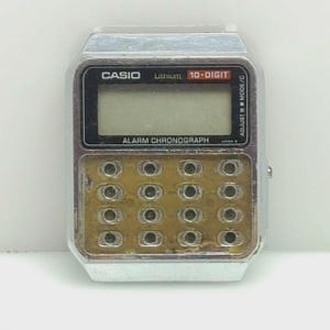 May include: A vintage Casio digital watch with a silver-tone case and a rectangular face. The watch features a calculator keypad, a digital display, and the text "CASIO", "Lithium", "10-DIGIT", and "ALARM CHRONOGRAPH".