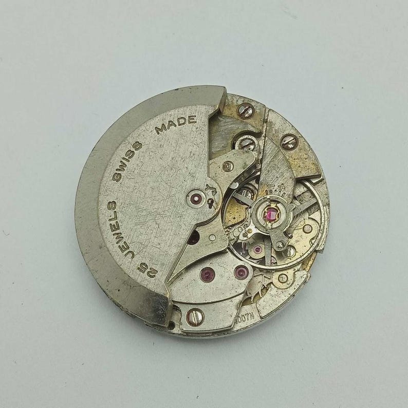 May include: A detailed view of a silver watch movement, showcasing its internal mechanisms. The words "SWISS MADE" and "25 JEWELS" are inscribed. The intricate design features small red jewels and various gears.