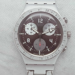 May include: A silver-toned Swatch chronograph watch with a dark brown face. The watch features white hands and markers, with sub-dials at the 2, 6, and 10 o'clock positions. The bezel has a tachymeter scale.