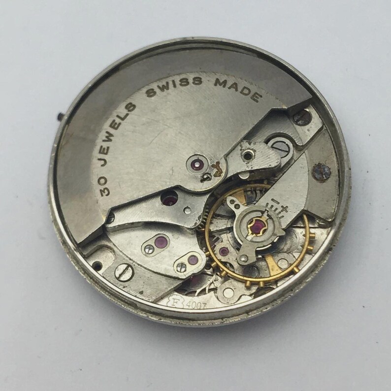 K&ouml;nnte beinhalten: Nahaufnahme eines Uhrwerks mit sichtbaren Zahnr&auml;dern, Juwelen und dem Text "SWISS MADE" und "30 JEWELS". Die Metallkomponenten sind silber- und goldfarben, mit roten Juwelen. Das Uhrwerk ist rund.