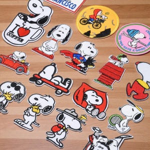 Op de afbeelding: Een verzameling Snoopy-patches met Snoopy in verschillende poses. Snoopy is te zien op een skateboard, als superheld of in zijn rode hondenhok. De patches zijn wit, zwart, rood en geel en hebben verschillende vormen.