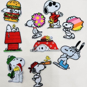 以下が含まれることがあります： 様々なポーズの、スヌーピーとウッドストックが描かれた刺繍パッチのコレクション。サングラスをかけたスヌーピー、ハンバーガー、赤い犬小屋の中のスヌーピー、望遠鏡を持ったスヌーピーなどのデザイン。パッチにはカラフルなアクセントが施されています。