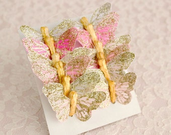 May include: A set of pink and gold glitter butterflies on a white card. The butterflies are arranged in a row and are attached to the card with wooden sticks.