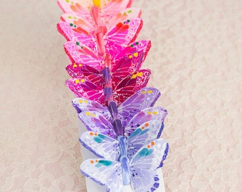 May include: A row of colorful paper butterflies in shades of pink, purple, and white. The butterflies are arranged in a gradient from pink to white.
