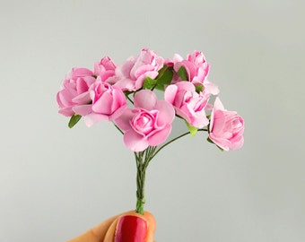 May include: A small bouquet of pink paper roses with green leaves and stems. The flowers are held in a hand with red nail polish against a light gray background. The roses are in full bloom.