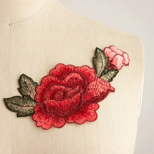 May include: A red embroidered rose with green leaves and a smaller pink rose on a beige fabric background.