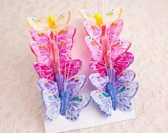 May include: Two sets of colorful butterfly decorations. Each set has three butterflies in shades of pink, purple, and blue. The butterflies have painted details and are attached to a white base with thin wire stems.