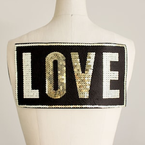 May include: Black rectangular sequin patch with the word "LOVE" in gold and white sequins. The patch is displayed on a mannequin. The sequins are sewn onto a black fabric, and the edges are outlined with gold sequins.