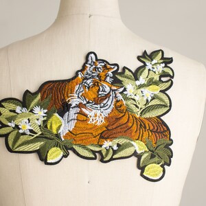 Pode incluir: Patch bordado com dois tigres com pelagem laranja e castanha, detalhes brancos e borda preta. Os tigres s&atilde;o rodeados por folhas verdes, flores brancas e frutos amarelos. Desenhado para vestu&aacute;rio.