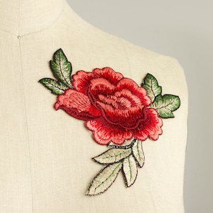 May include: A red rose embroidered patch featuring detailed stitching with layered petals in shades of crimson and dark red, accompanied by green leaves with visible thread texture, displayed on a neutral-colored fabric surface.