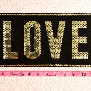 Może przedstawiać: Czarna prostokątna naszywka z napisem "LOVE" wyszytym złotymi cekinami. Naszywka ma około 25 cm długości i 13 cm szerokości. Tło stanowi jasnor&oacute;żowa koronka.