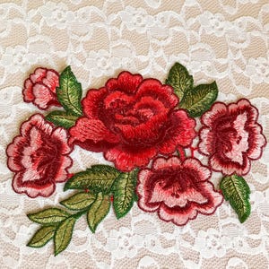 May include: Embroidered red roses with green leaves on a white lace background. The roses are in a cluster and have a 3D effect.