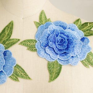 May include: Two embroidered floral appliques featuring layered blue petals in varying shades of light and dark blue, accented by detailed green leaves with visible stitching, positioned on a neutral-colored fabric surface.