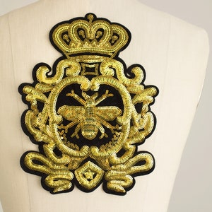 May include: A gold sequined patch with a black backing. The patch features a bee design within a decorative frame, topped with a crown. The patch is approximately 6 inches wide and 7 inches tall.