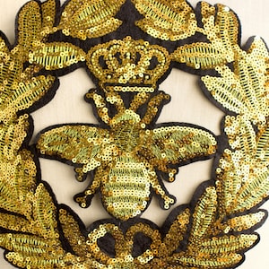 May include: A gold sequined bee patch with a crown and laurel wreath design. The bee is the focal point, surrounded by a circular wreath. The patch is made with gold sequins on a black felt backing, creating a luxurious and detailed embellishment.