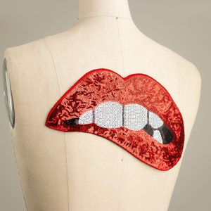 Pode incluir: Um patch de l&aacute;bios vermelhos com lantejoulas, dentes brancos e uma marca de mordida preta, exibido em um manequim. O patch &eacute; um acess&oacute;rio vibrante, perfeito para adicionar um toque de glamour.
