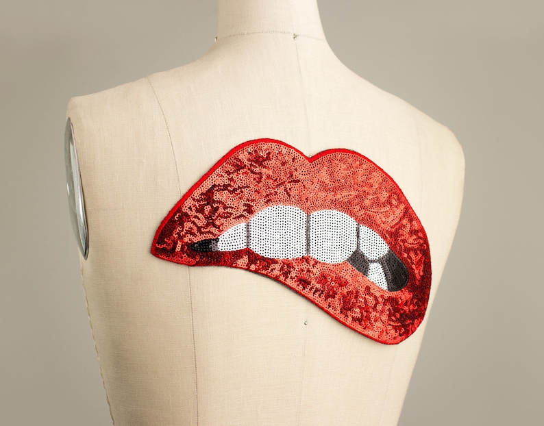 Pode incluir: Um patch de l&aacute;bios vermelhos com lantejoulas, dentes brancos e uma marca de mordida preta, exibido em um manequim. O patch &eacute; um acess&oacute;rio vibrante, perfeito para adicionar um toque de glamour.