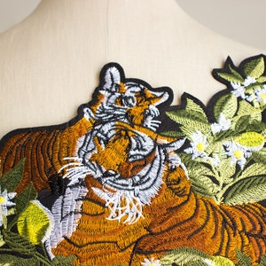 May include: Embroidered iron-on patch featuring a tiger curled up in a bed of green leaves with white flowers and yellow fruit.
