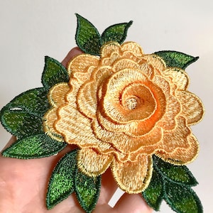 May include: A yellow embroidered flower with green leaves. The flower is made of multiple layers of fabric and has a spiral design in the center.