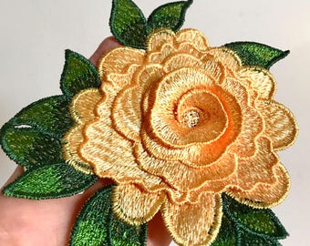 May include: A yellow embroidered flower with green leaves. The flower is made of multiple layers of fabric and has a spiral design in the center.