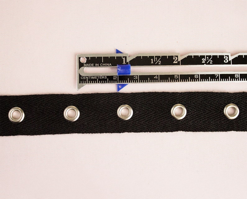 Pu&ograve; includere: Nastrino in cotone nero con quattro occhielli argentati. Il nastro &egrave; largo 2,5 cm e viene misurato con un righello.