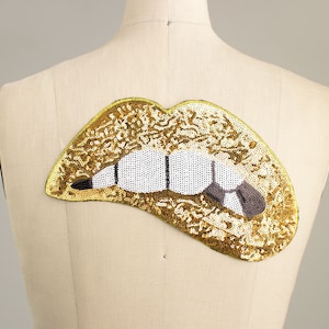 May include: A gold sequined lips patch with white and black detailing for the teeth and a gray tongue. The patch is attached to a mannequin. The lips are outlined in a thin yellow border.