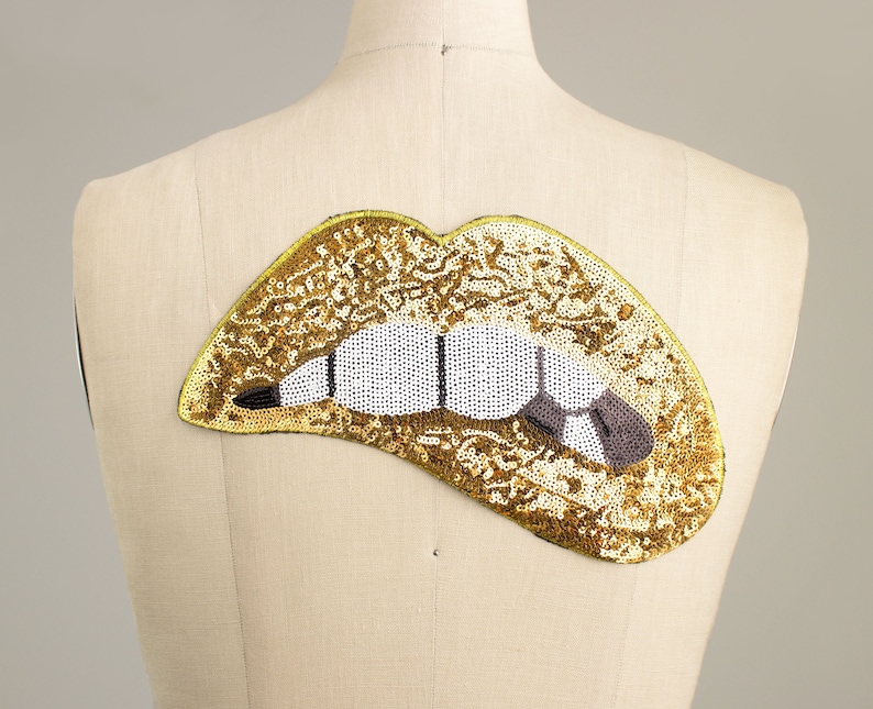 Pode incluir: Um patch de l&aacute;bios com lantejoulas douradas com detalhes brancos e pretos para os dentes e uma l&iacute;ngua cinza. O patch est&aacute; preso a um manequim. Os l&aacute;bios s&atilde;o delineados por uma fina borda amarela.