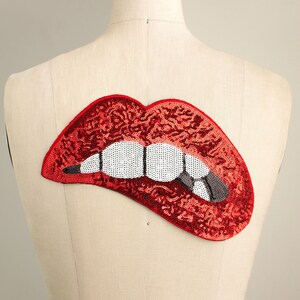 May include: A red sequined lips patch with white teeth and a gray tooth biting the lower lip. The patch is attached to a mannequin. The lips are a bold, eye-catching design, perfect for adding flair to clothing or accessories.