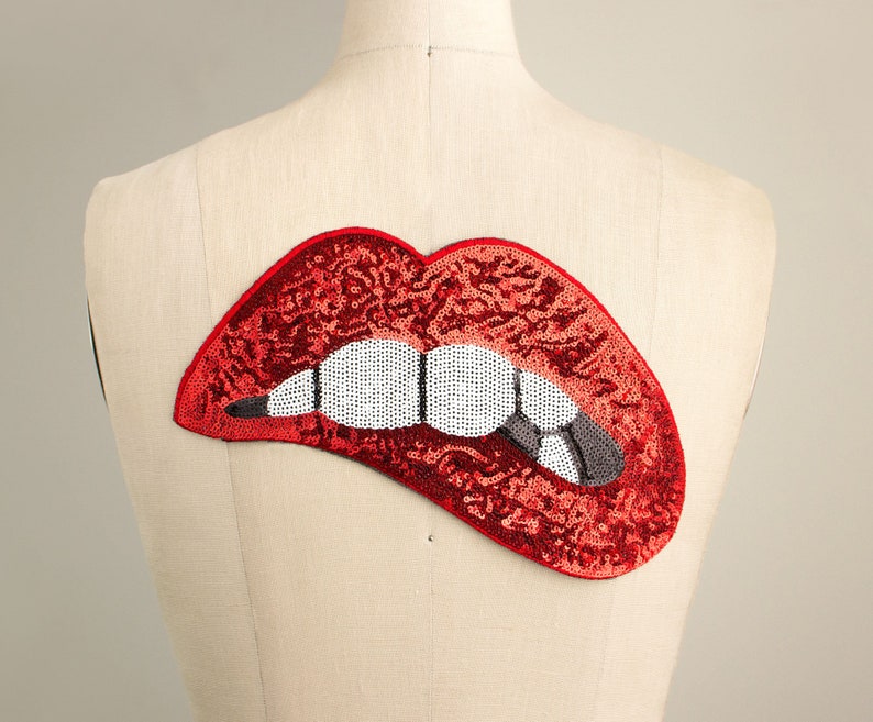 Pode incluir: Um patch de l&aacute;bios vermelhos com lantejoulas, com dentes brancos e um dente cinza mordendo o l&aacute;bio inferior. O patch est&aacute; preso a um manequim. Os l&aacute;bios s&atilde;o um design arrojado, perfeito para adicionar estilo a roupas ou acess&oacute;rios.