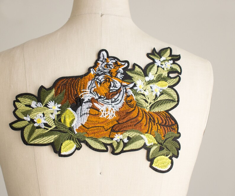 Pode incluir: Patch bordado com dois tigres com pelagem laranja e castanha, detalhes brancos e borda preta. Os tigres s&atilde;o rodeados por folhas verdes, flores brancas e frutos amarelos. Desenhado para vestu&aacute;rio.
