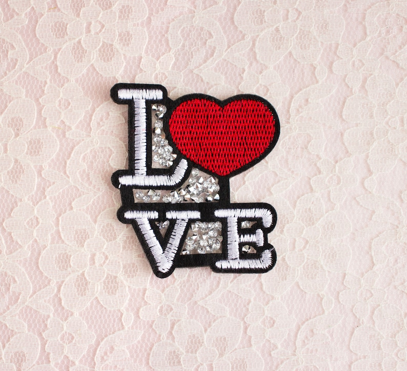 Może przedstawiać: Haftowana naszywka z napisem "LOVE" w kolorze białym, obramowana na czarno. "O" to czerwone serce. Litery wypełnione są srebrnymi kryształkami. Do odzieży lub akcesori&oacute;w.