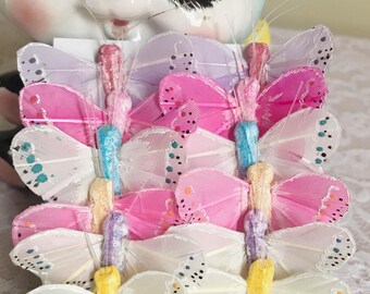 May include: A set of 10 colorful butterfly embellishments. The butterflies are made of feathers and have a glittery finish. They are arranged in a stack, with the white butterflies at the bottom and the pink, purple, and blue butterflies on top.
