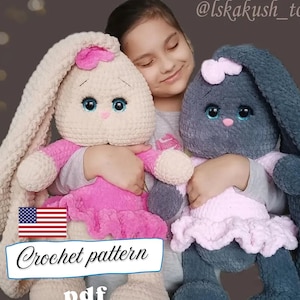 May include: Two plush crochet bunny dolls, one cream and one grey, are held by a person. The bunnies have large eyes, pink accents, and wear pink dresses. The image includes text that says "Crochet pattern pdf".