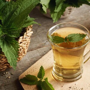 May include: A clear glass mug filled with light amber herbal tea, garnished with a fresh green nettle leaf floating on the surface. Beside the mug sits a woven wicker basket overflowing with vibrant green nettle leaves and stems.