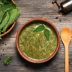 May include: A round brown ceramic bowl filled with green nettle soup, garnished with two fresh green nettle leaves on the surface. Beside the bowl sits a wooden spoon, a small basket of fresh nettle leaves, and a metal pepper mill on a wooden surface.
