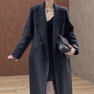 May include: A dark gray double-breasted coat with a notched collar and two rows of buttons. The coat has a tailored fit and a long length. A black handbag is held in the hand. The coat is a stylish outerwear garment.