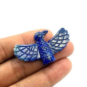 May include: A carved lapis lazuli bird figurine with outstretched wings. The bird is a deep blue color with white accents on the wings. The figurine is held in a hand against a white background.