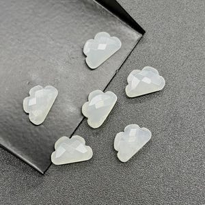 May include: Six white, cloud-shaped gemstone beads with a faceted design are displayed on a dark grey surface. The beads are translucent and catch the light. These beads are likely for jewellery making or craft projects.