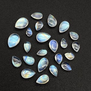 May include: A collection of pear-shaped moonstone gemstones with a translucent, iridescent quality. The stones display a range of blues, whites, and yellows, arranged on a dark surface. These polished stones are ideal for jewelry making.