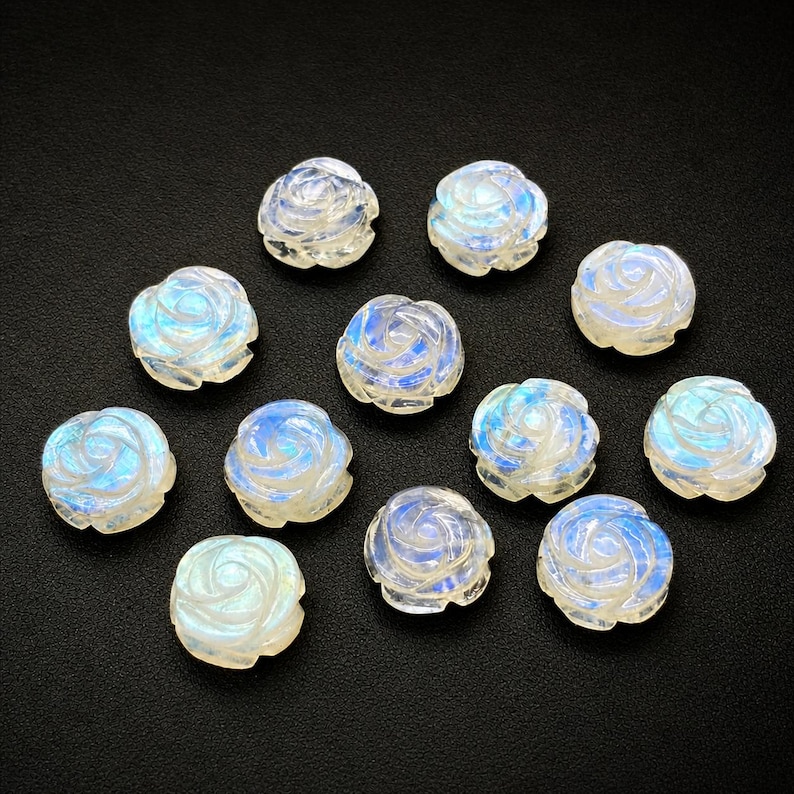Puede incluir: Una colecci&oacute;n de cuentas de rosa de piedra lunar talladas. Cada cuenta est&aacute; esculpida en forma de rosa, mostrando una gama de colores que van del blanco al azul. Las cuentas est&aacute;n dispuestas sobre una superficie negra, mostrando sus intrincados detalles.