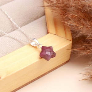 May include: A silver necklace featuring a star-shaped pendant. The pendant is a faceted, deep pink gemstone. The necklace is displayed in a wooden box with a light-coloured interior, and dried ornamental grass.
