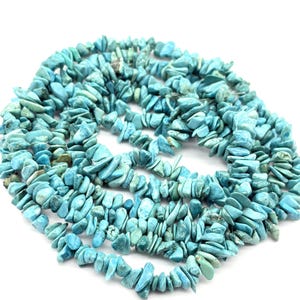 Puede incluir: Cadenas de cuentas de virutas turquesas en varios tonos de azul y verde. Las formas irregulares de las cuentas crean una apariencia texturizada. Las cuentas están esparcidas sobre una superficie blanca, ideales para la creación de joyas.