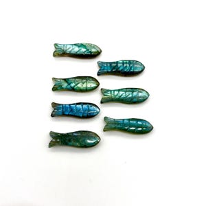 Puede incluir: Siete figuras de peces talladas en tonos azules y verdes. Cada pez tiene aletas y escamas detalladas, con variaciones de color y textura. Las figuras están dispuestas sobre un fondo blanco, mostrando su tema acuático.