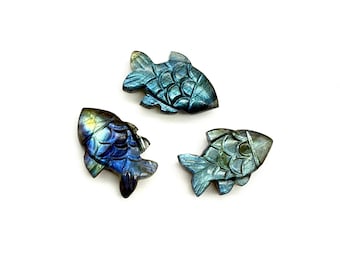 Puede incluir: Tres figuras de peces talladas en tonos azules, verdes y turquesas. Los peces tienen patrones de escamas detallados y están dispuestos sobre un fondo blanco. Estos artículos decorativos podrían usarse como colgantes o amuletos.