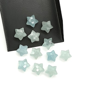 Pode incluir: Uma coleção de pedras preciosas em forma de estrela facetadas, na cor água-marinha. As pedras são exibidas em uma superfície preta e um fundo branco. As estrelas têm uma qualidade translúcida, com facetas internas visíveis.