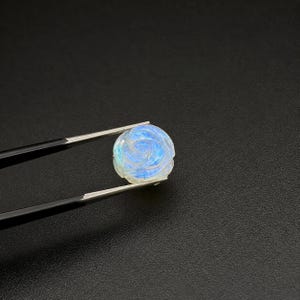 Puede incluir: Una gema redonda tallada, sujeta con pinzas. La gema es blanca con reflejos azules e iridiscentes, y est&aacute; tallada con un patr&oacute;n de rosa. El fondo es negro.