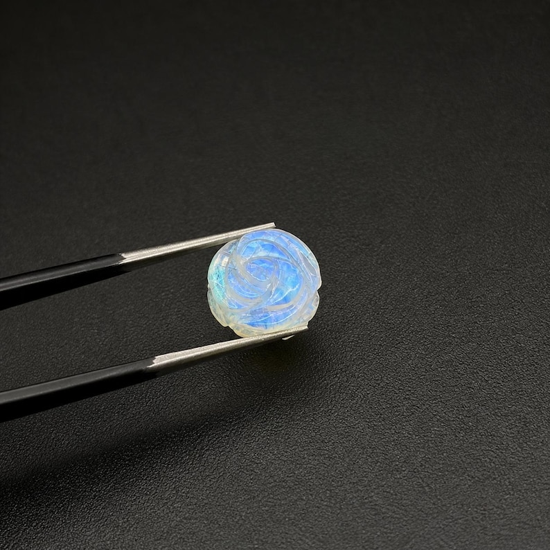 Puede incluir: Una gema redonda tallada, sujeta con pinzas. La gema es blanca con reflejos azules e iridiscentes, y est&aacute; tallada con un patr&oacute;n de rosa. El fondo es negro.