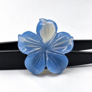 May include: A translucent light blue flower-shaped carving with five rounded petals. The surface features delicate, radiating carved lines that originate from the center, creating a subtle depth and a smooth, polished finish on the gemstone.