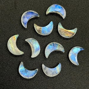 Pode incluir: Nove pedras preciosas iridescentes em forma de lua crescente, com tons azuis, amarelos e brancos. As pedras polidas estão dispostas sobre uma superfície escura, mostrando suas variações de cor e qualidades reflexivas únicas. Ideal para joalheria.
