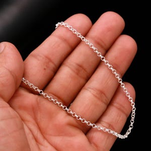 May include: A delicate silver chain necklace with a classic cable link design. The necklace is displayed against a dark background, highlighting its bright, reflective surface. The chain is approximately 40 cm long.